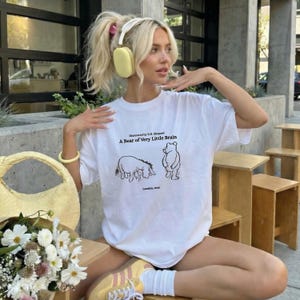 May include: A white short-sleeve t-shirt featuring a black line drawing of Eeyore and Winnie the Pooh. The text reads: Illustrated by E.H. Shepard, A Bear of Very Little Brain, London, 1926. The shirt has a crew neckline and a relaxed fit.