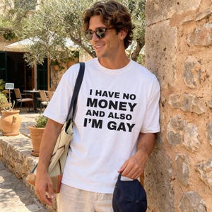 Könnte beinhalten: Weißes T-Shirt mit dem Text "I HAVE NO MONEY AND ALSO I'M GAY" in Schwarz. Die Person trägt eine Sonnenbrille und hält eine schwarze Kappe. Eine beigefarbene Tragetasche hängt über der Schulter.