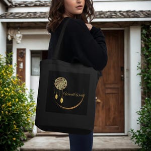 Op de afbeelding: Een zwarte tote bag met een goudkleurige afbeelding van een cirkelvormig mandalapatroon met hangende lantaarnachtige ornamenten en een halve maan. De tekst WardaWorld is in een gouden scriptlettertype in het midden van het ontwerp gedrukt.