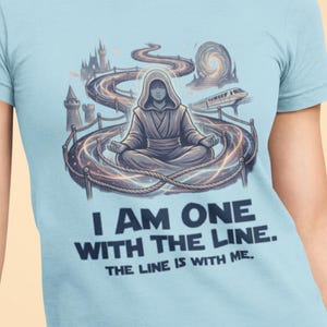 Puede incluir: Camiseta azul claro con un gráfico de una figura con túnica meditando dentro de un diseño circular, con un camino sinuoso y un castillo en el fondo. El texto "I AM ONE WITH THE LINE. THE LINE IS WITH ME." está impreso debajo.
