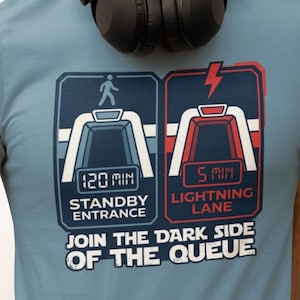 May include: A light blue t-shirt with a graphic featuring two stylized ride entrances, one labeled "Standby Entrance" with a 120-minute wait time and the other "Lightning Lane" with a 5-minute wait. The text "Join the Dark Side of the Queue" is below.