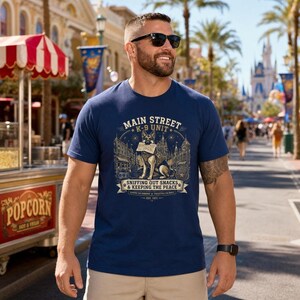 May include: A navy blue short-sleeve t-shirt featuring a graphic print of a German Shepherd dog holding a snack. The text reads MAIN STREET K-9 UNIT, SNIFFING OUT SNACKS & KEEPING THE PEACE, GUARDING THE KINGDOM, PROTECTING THE MAGIC, and EST. 1971.