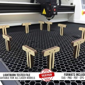 Op de afbeelding: Een verzameling T-vormige houten stukken, waarschijnlijk voor een laser-snijproject, gerangschikt op een honingraatoppervlak. De stukken hebben een lichte houtkleur met donkerbruine accenten. De afbeelding bevat tekst die aangeeft dat het bestand is getest.