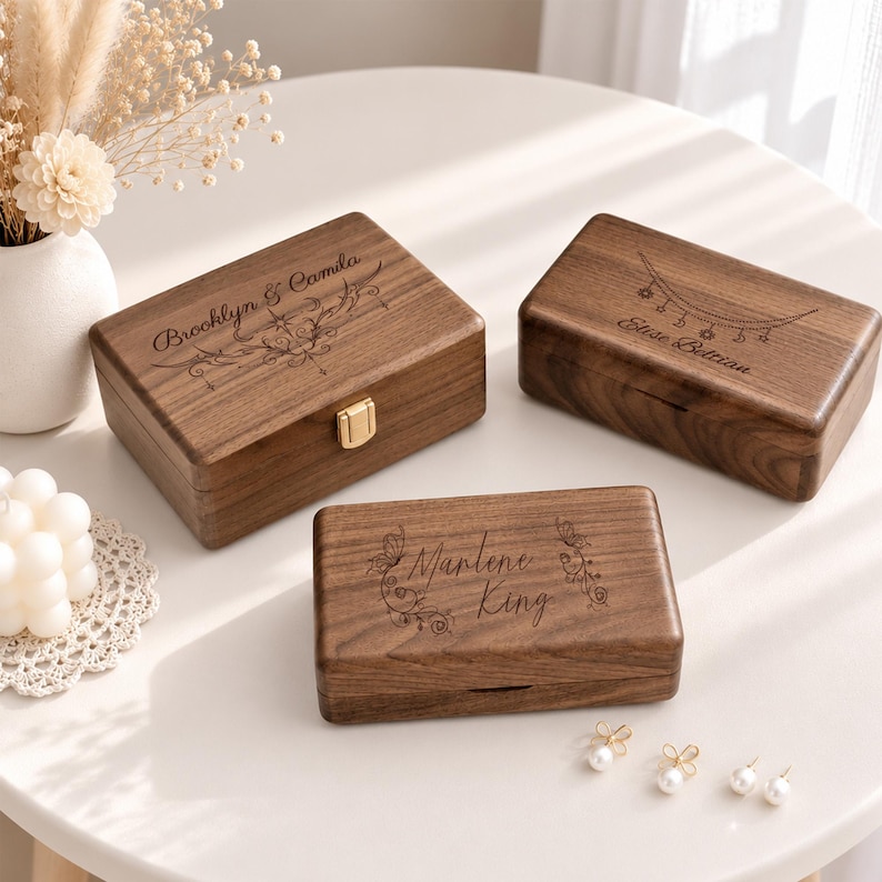 Pu&ograve; includere: Tre portagioie rettangolari in legno marrone scuro con coperchi incernierati. Ogni scatola presenta testo inciso al laser e motivi decorativi sulla superficie superiore. Una scatola ha una chiusura in metallo dorato, le altre sono lisce.