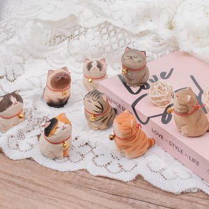 May include: A collection of carved wooden cat figurines in various colors and patterns. Each cat wears a red collar with a small gold bell. The cats are arranged on a white lace surface, with a pink book and decorative balls in the background.