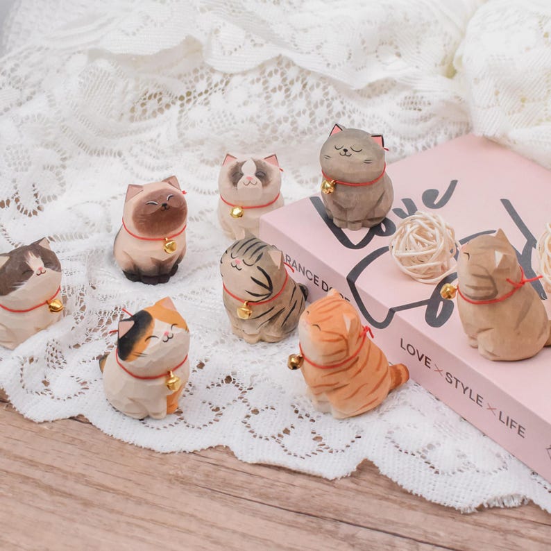 May include: A collection of carved wooden cat figurines in various colors and patterns. Each cat wears a red collar with a small gold bell. The cats are arranged on a white lace surface, with a pink book and decorative balls in the background.