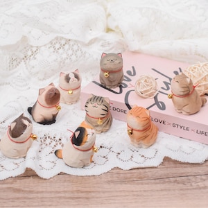 May include: A collection of wooden cat figurines in various poses and colors, each wearing a red collar with a small gold bell. The cats are painted in shades of brown, gray, orange, and white. The figurines are displayed on a white lace surface.