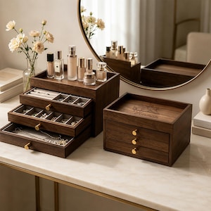 Può includere: Due organizer in legno scuro con pomelli dorati. L'unità a sinistra presenta tre cassetti estraibili con rivestimento in velluto grigio contenenti gioielli, sormontati da flaconi di profumo. L'unità a destra ha tre cassetti e il nome "Herman" inciso.