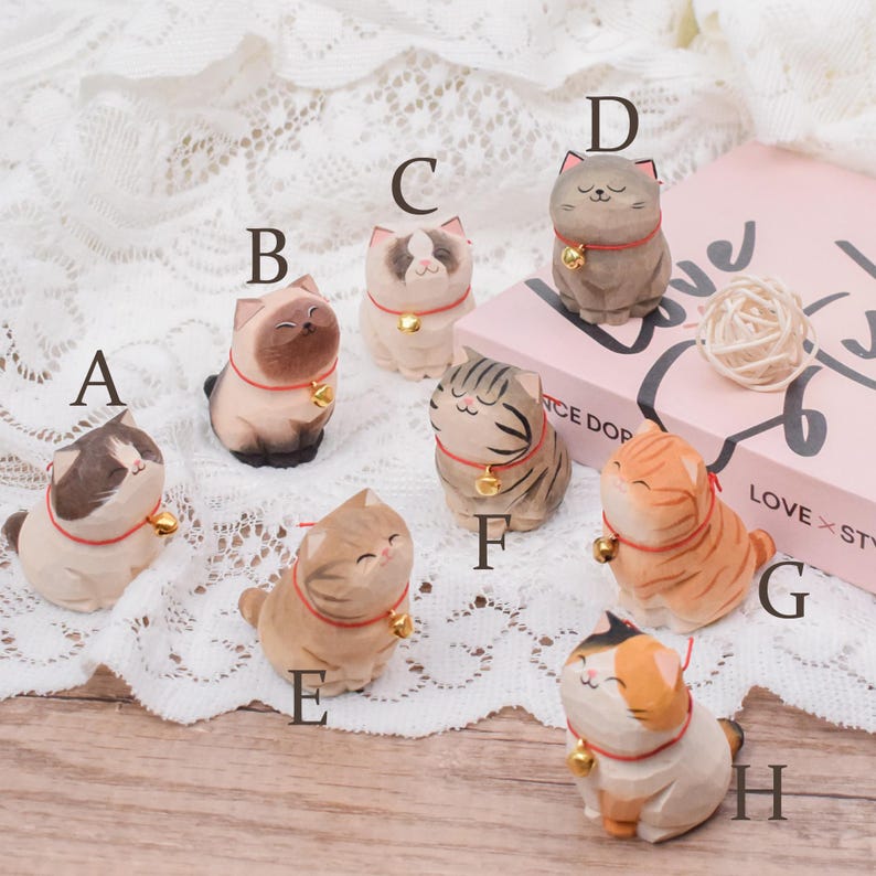 May include: A collection of hand-carved wooden cat figurines in various colors and patterns. Each cat wears a red collar with a small gold bell. The cats have different facial expressions and poses, adding to their charm.