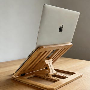 Puede incluir: Un soporte para portátil de bambú natural con un portátil encima. El soporte está inclinado para un uso ergonómico y presenta el texto "Better than yesterday". El portátil tiene un acabado plateado y el logotipo de Apple.