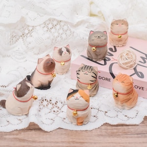 May include: A collection of handcrafted wooden cat figurines in various colors and patterns. Each cat wears a red collar with a small gold bell. The cats are arranged on a white lace surface, with a book and a ball of twine in the background.