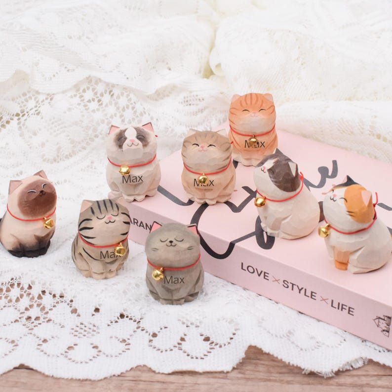 May include: A collection of hand-carved wooden cat figurines in various colors and patterns. Each cat has a red collar with a gold bell and the name "Max" engraved. The figurines are arranged on a pink book with the text "LOVE STYLE LIFE".