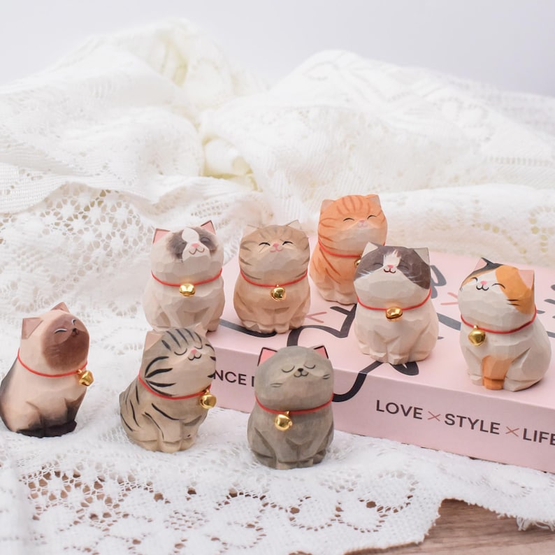 May include: A collection of nine carved wooden cat figurines. Each cat has a unique color pattern, a small bell collar, and a smiling expression. The cats are arranged on a pink box with the words "LOVE STYLE LIFE".