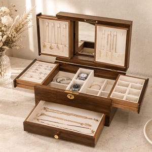 May include: A dark wood jewelry box with a hinged lid, a central mirror, and multiple compartments. The interior features cream-colored lining, hooks for necklaces, ring slots, and a pull-out drawer containing gold chains and various jewelry pieces.