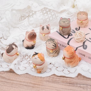 May include: A collection of hand-carved wooden cat figurines in various colors and patterns. Each cat has a red collar with a gold bell. The cats are arranged on a white lace surface, with a pink box and a ball of twine in the background.