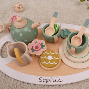 May include: A wooden tea set with a green polka-dot teapot, cups, saucers, spoons, and a serving tray. The set includes a flower-shaped cookie and a lemon slice. The tray is inscribed with the name "Sophia".