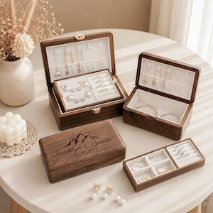 Puede incluir: Tres joyeros rectangulares de madera con interiores de terciopelo blanco. Una caja está cerrada y grabada con el texto Brooklyn & Camila 09.19.2025 y un gráfico de montaña. Las cajas abiertas contienen varios pendientes, collares y anillos de oro.