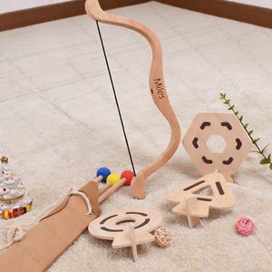 May include: A wooden toy bow and arrow set with a quiver, target, and other wooden shapes. The bow has the name "Miles" printed on it. The arrows have blue, yellow, and red tips. The set is designed for children.