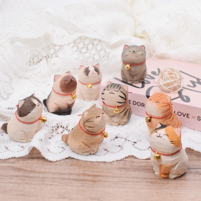 May include: A collection of hand-carved wooden cat figurines in various colors and patterns. Each cat wears a red collar with a gold bell. The cats are arranged on a wooden surface with a white lace cloth in the background.