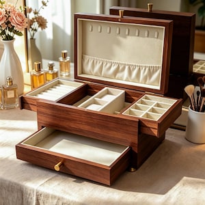 May include: A dark wood jewelry box with a hinged lid, a pull-out drawer, and a cream-colored interior. The lid features necklace hooks and a pocket, while the interior contains multiple compartments, including ring rolls and small square dividers.