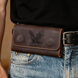 Op de afbeelding: Een donkerbruine leren riemtas met een klepsluiting, voorzien van een in laser gegraveerde adelaarsafbeelding en de naam MICHAEL eronder gecentreerd. Het rechthoekige accessoire zit vast aan een zwarte leren riem over een blauwe spijkerbroek.