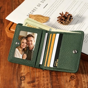 Puede incluir: Una cartera plegable de color verde bosque con acabado de cuero texturizado, que incluye una ventana transparente con la fotografía de una pareja, múltiples ranuras para tarjetas y un cierre de botón a presión metálico, mostrada abierta.
