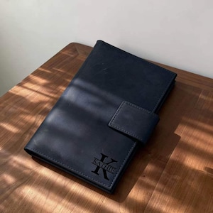 以下が含まれることがあります： フラップ開閉式の濃い青色の革製ジャーナル。ジャーナルには、前面に文字「K」と名前「Kathie」が刻印されています。木製の表面に置かれています。