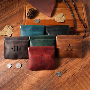 May include: A collection of leather coin pouches in various colors, including brown, teal, black, green, and red. Each pouch features a metal clasp and personalized initials or a cross design. The pouches are displayed on a wooden surface.