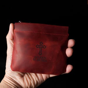 May include: A red leather pouch with a zipper closure. The pouch features a cross design and the name "AILEEN" with the date "02.05.2026" engraved on the front. The pouch is held in a hand against a black background.