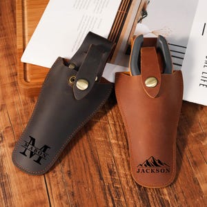 May include: Two leather tool holsters with snap closures. The dark brown holster features a black monogram with the text MABON. The light brown holster contains a tool with black handles and features a black mountain graphic with the text JACKSON.