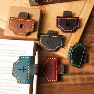 Puede incluir: Una colección de sujetapáginas de cuero en varios colores, incluyendo marrón, verde azulado y burdeos. Cada sujetapáginas presenta un diseño único, como una araña, un búho, una cruz y el nombre "Aileen". Los bordes están cosidos.