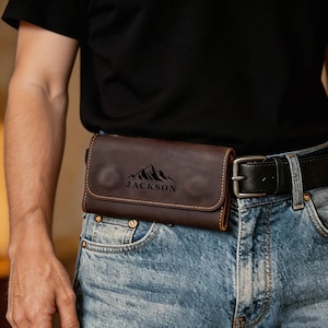 May include: A dark brown leather belt pouch featuring a flap closure with visible stitching along the edges. The front of the pouch is engraved with a mountain range graphic above the name JACKSON. It is attached to a black leather belt.