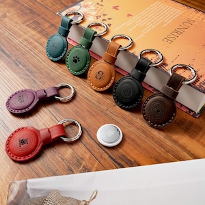 May include: A selection of leather keyrings in shades of teal, green, brown, black, purple, and red. Each keyring has a silver ring and an embossed design, including a paw print, initials, or a cat. The keyrings are on a wooden surface.