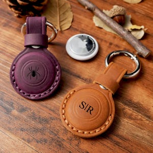 May include: Two leather AirTag holders with key rings. One is burgundy with a spider graphic, the other is brown with the initials "S/R". A white AirTag is also visible. The holders are on a wooden surface with autumn leaves.