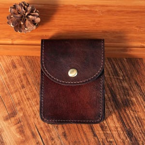 Puede incluir: Una funda rectangular de cuero marrón oscuro con una solapa redondeada asegurada por un solo botón de presión metálico de color latón. El cuero presenta una textura granulada visible y bordes bruñidos más oscuros, con costuras perimetrales.
