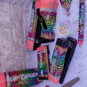 May include: A collection of school supplies, including a pencil, stapler, pen, and scissors, all decorated with a rainbow zebra print and the name "Ms. Thomas". The items have a glittery finish and neon pink accents.