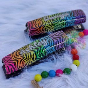 May include: Two staplers with rainbow zebra print and the names "Ashley" and "Danya" in cursive. A beaded pacifier clip with colorful beads and a jute cord is also present. The background is a fluffy white surface.