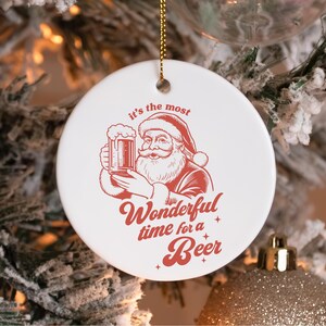 May include: A round white ceramic ornament featuring a red graphic of Santa Claus holding a beer mug. The text reads, "it's the most Wonderful time for a Beer" in red lettering. The ornament hangs from a gold string against a blurred tree background.