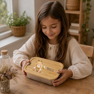 May include: A rectangular metal lunch box with a light-colored bamboo lid. The lid features a printed illustration of a cartoon bear dressed as a bee and the name AMALIA in black text. The lid is secured with two silver-colored metal latches.