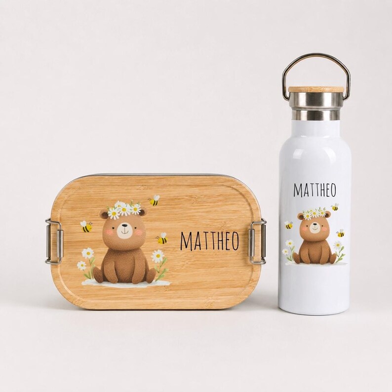 May include: A rectangular bamboo lunch box and a white metal water bottle. Both items feature a brown bear illustration wearing a daisy crown, surrounded by small bees and the name MATTHEO printed in black, sans-serif capital letters.