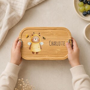 May include: A rectangular bamboo lunch box with rounded corners and metal side latches. The lid features a printed illustration of a brown bear wearing a yellow striped bee costume with wings, two small bees, and the name Charlotte in black text.