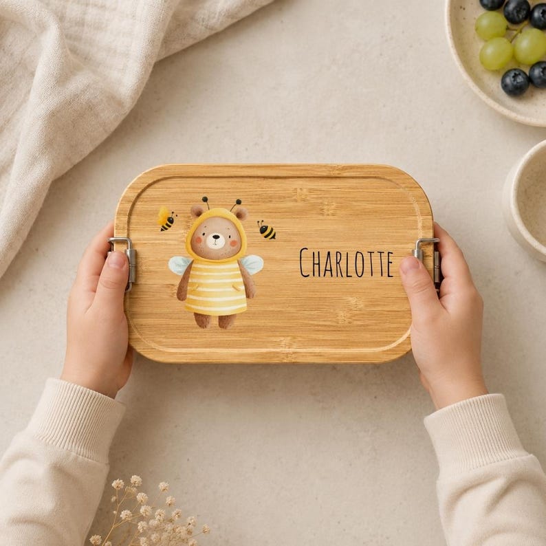 May include: A rectangular bamboo lunch box with rounded corners and metal side latches. The lid features a printed illustration of a brown bear wearing a yellow striped bee costume with wings, two small bees, and the name Charlotte in black text.