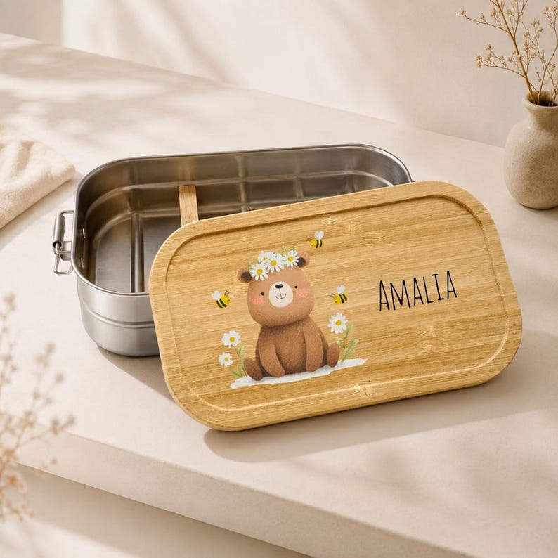May include: A rectangular stainless steel lunch box with a removable bamboo lid. The lid features a printed illustration of a brown bear wearing a daisy crown, surrounded by small bees and flowers, with the name AMALIA printed in black text.