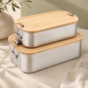 May include: Two rectangular food containers stacked on top of each other. Each container features a brushed silver metal base with rounded corners and a light-colored bamboo wood lid. Metal wire latches secure the lids to the containers.