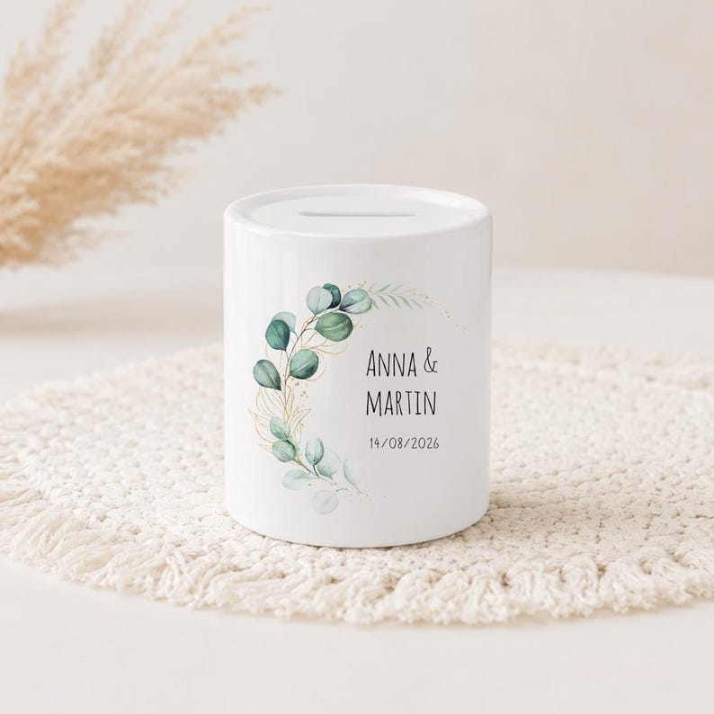 May include: A white ceramic money box featuring a printed design of green eucalyptus leaves and gold-coloured stems. The text reads ANNA & MARTIN followed by the date 14/08/2026. The cylindrical container has a horizontal coin slot on the top surface.
