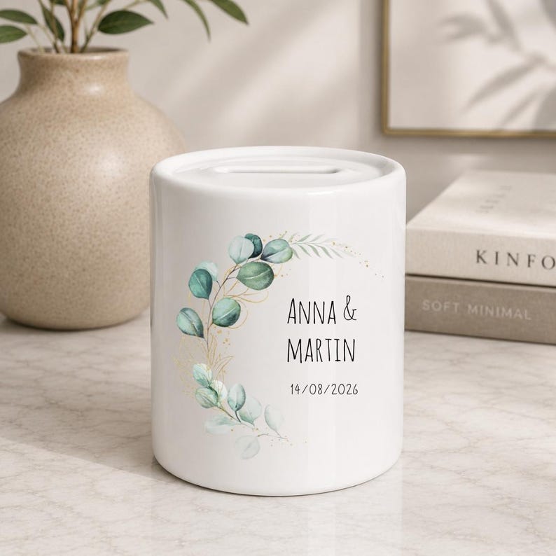 May include: A white ceramic money box featuring a slot on top and a printed design of green eucalyptus leaves with gold accents. The text reads ANNA & MARTIN and the date 14/08/2026 in a simple black sans-serif font on the front surface.