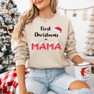 May include: A beige crewneck sweatshirt featuring the black text First Christmas as and the red text MAMA with white snow accents on the letters. A small red and white Santa hat graphic is positioned above the word Christmas on the front.