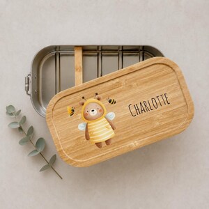 May include: A rectangular stainless steel lunch box with a removable bamboo lid. The lid features a printed illustration of a brown bear wearing a yellow and white striped bee costume with wings, surrounded by small bees and the name CHARLOTTE.