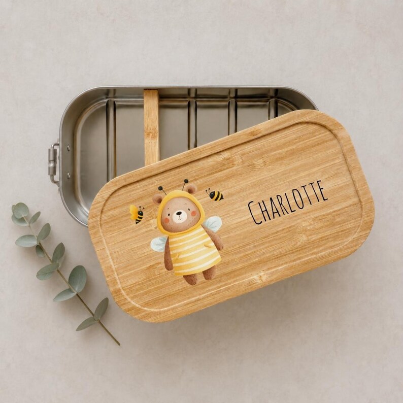 May include: A rectangular stainless steel lunch box with a removable bamboo lid. The lid features a printed illustration of a brown bear wearing a yellow and white striped bee costume with wings, surrounded by small bees and the name CHARLOTTE.
