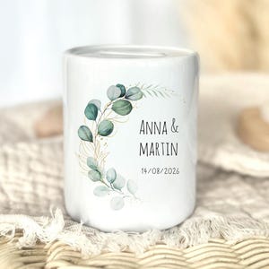 May include: A white ceramic money box featuring a curved eucalyptus branch design in shades of green and gold. The text ANNA & MARTIN is printed in black, followed by the date 14/08/2026. The cylindrical container has a coin slot on top.